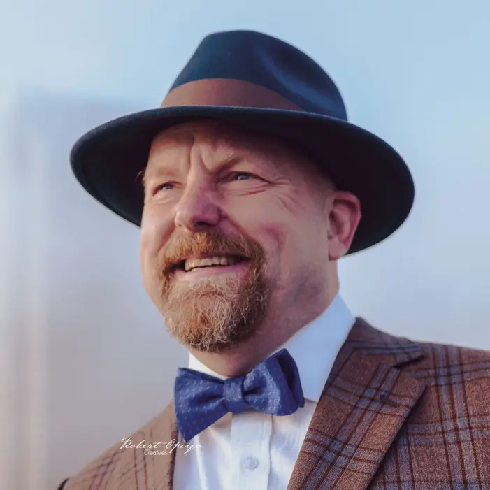 Kent looking up & smiling wearing a blue fedora & a bow tie. Photo credit: Robert Opiyo