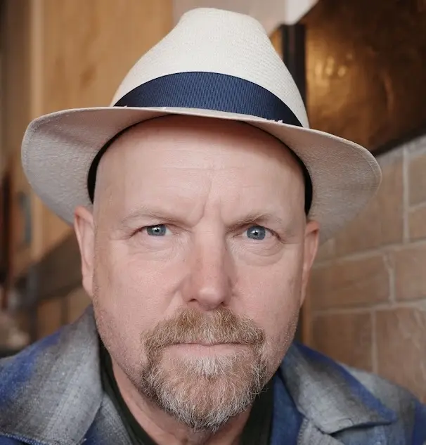 Kent looking directly at the camera in a white straw hat & blue plaid shirt. Photo credit: Kunal Bhalla